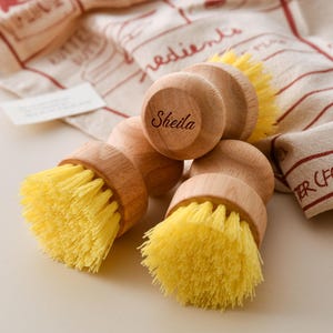 Puede incluir: Tres cepillos de fregar de madera con cerdas amarillas. La parte superior de un cepillo tiene el nombre "Sheila" grabado.