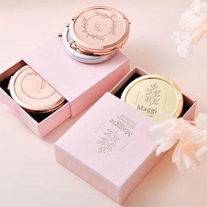 May include: Two pink boxes containing gold and rose gold compact mirrors. The mirrors feature engraved floral designs and the text "Maggi" and "Bride Tribe" on them.