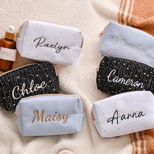 May include: A collection of personalized cosmetic bags in various colors, including white, black, and blue, each adorned with a name in elegant script. The bags feature a gold zipper and a starry pattern. A brown bottle with a pump is also present.
