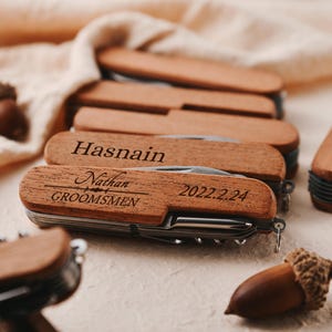 May include: A set of personalized wooden pocket knives with engraved names and the date "2022.2.24". The knives have a brown wooden handle and silver blades. The text on the knives reads "Hasnain" and "Nathan GROOMSMEN".