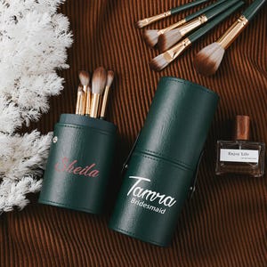 May include: Two green makeup brush holders with gold lids. The holder on the left says "Sheila" and the holder on the right says "Tamra Bridesmaid".