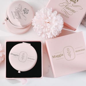 以下が含まれることがあります： 2つのピンク色のコンパクトミラーに、パーソナライズされた彫刻が施されています。1つのミラーには、花柄のデザインと、Maggieという名前と2020年5月5日の日付が刻まれています。もう1つのミラーには、王冠のデザインと、Naomiという名前と2020年5月5日の日付が刻まれています。