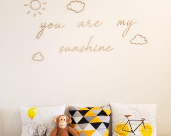 May include: A nursery scene with wooden sun and cloud decorations, and the phrase "you are my sunshine" on a cream wall. Below are three throw pillows with animal and geometric designs, and soft toys.