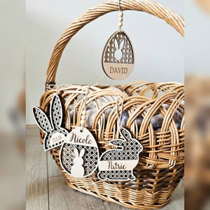 Può includere: Un cestino pasquale in vimini intrecciato con etichette di legno a forma di uova e coniglietti pasquali. Le etichette sono tagliate al laser con nomi come "David", "Nicole", "Julia" e "Patric". Le etichette sono attaccate al cestino con spago.