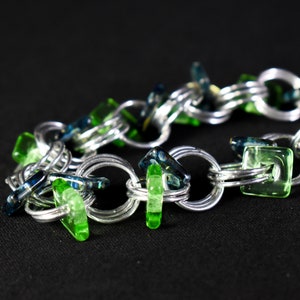 May include: A silver chain bracelet with green and blue glass details. The bracelet is made of interconnected silver links, with bright green and blue glass elements. The glass pieces are in different shapes, including squares and oblongs.