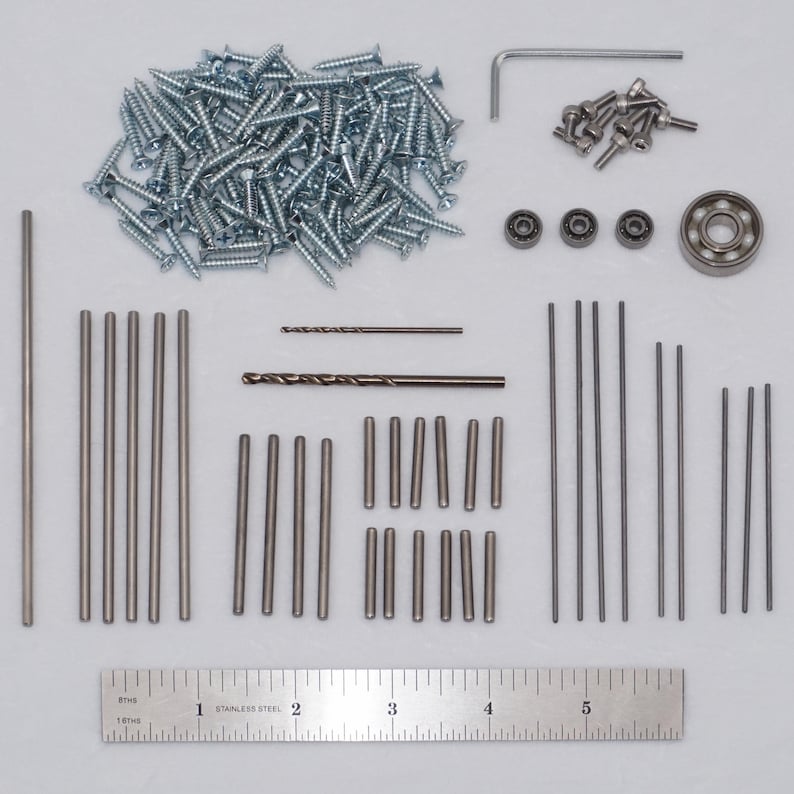 May include: A hardware kit containing a large pile of silver screws, a small L-shaped hex key, several small bolts, four ball bearings, and various metal rods and drill bits of different lengths. A 6-inch stainless steel ruler is placed at the bottom.
