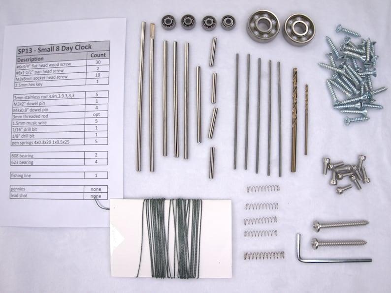 K&ouml;nnte beinhalten: Ein Bausatz f&uuml;r eine kleine 8-Tage-Uhr. Der Bausatz enth&auml;lt verschiedene Metallteile, Schrauben, Federn und eine kleine Spule mit Schnur. Die Teile liegen auf einer wei&szlig;en Oberfl&auml;che. Der Bausatz enth&auml;lt auch eine Liste der Teile und ihrer Mengen.