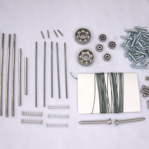 Op de afbeelding: Assortiment van metalen hardware, waaronder schroeven, veren, staven en lagers. De hardware is gerangschikt op een wit oppervlak.