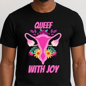 May include: Black t-shirt featuring a pink uterus graphic with floral accents and the text "QUEEF WITH JOY" in pink. The design includes yellow and pink flowers, and purple leaves.