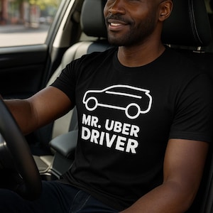 May include: Black t-shirt with a white outline of a car and the text "MR. UBER DRIVER". The shirt is worn by a person sitting in the driver's seat of a car.