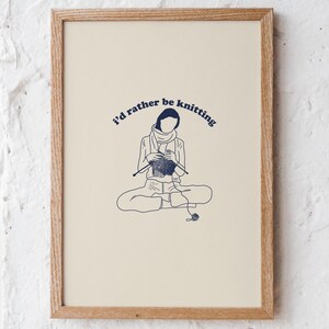 Pu&ograve; includere: Una stampa incorniciata con uno sfondo crema e un'illustrazione blu di una persona seduta a gambe incrociate e che fa la maglia. Il testo "I'd rather be knitting" &egrave; scritto in un arco blu sopra la persona.