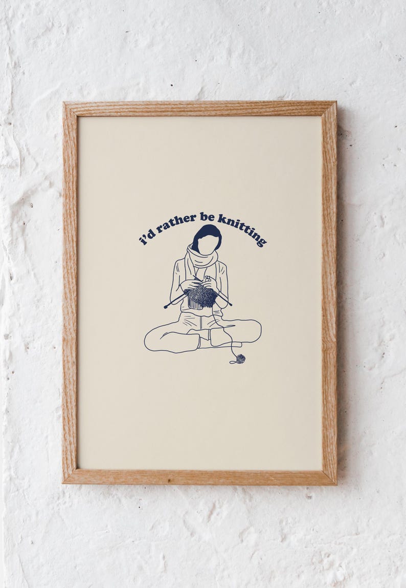Pu&ograve; includere: Una stampa incorniciata con uno sfondo crema e un'illustrazione blu di una persona seduta a gambe incrociate e che fa la maglia. Il testo "I'd rather be knitting" &egrave; scritto in un arco blu sopra la persona.