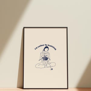 Pu&ograve; includere: Una stampa incorniciata nera con sfondo bianco e un'illustrazione blu di una persona che fa la maglia. Il testo "I'd rather be knitting" &egrave; scritto in blu in corsivo.