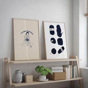 Pu&ograve; includere: Due stampe incorniciate su uno scaffale di legno. La stampa a sinistra presenta un disegno a linea di una persona che fa la maglia con il testo "I'd rather be knitting". La stampa a destra presenta una collezione di gomitoli di lana blu.