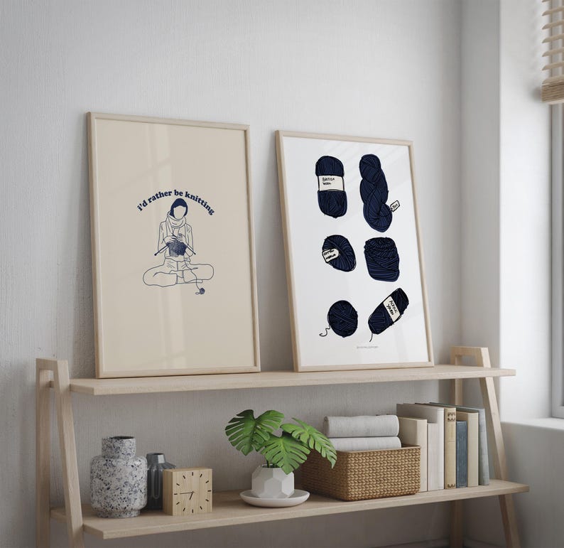 Pu&ograve; includere: Due stampe incorniciate su uno scaffale di legno. La stampa a sinistra presenta un disegno a linea di una persona che fa la maglia con il testo "I'd rather be knitting". La stampa a destra presenta una collezione di gomitoli di lana blu.