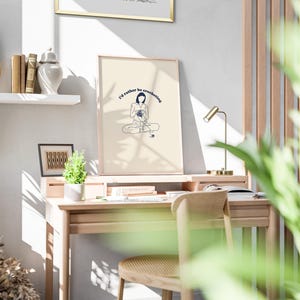 Pu&ograve; includere: Una stampa incorniciata con uno sfondo crema e un disegno a linea blu di una persona seduta con il testo "I'd rather be crocheting". La stampa &egrave; appesa a una parete bianca sopra una scrivania di legno con una lampada e una pianta.