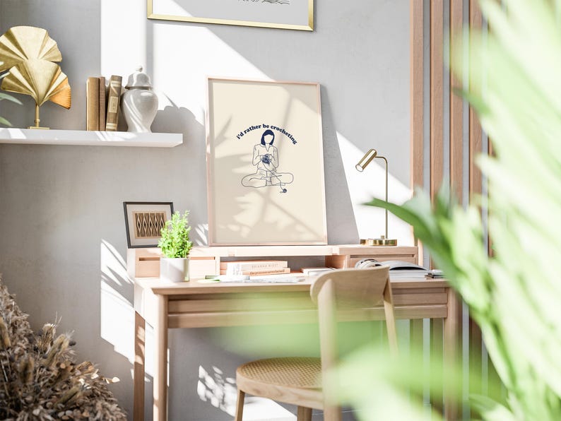 Pu&ograve; includere: Una stampa incorniciata con uno sfondo crema e un disegno a linea blu di una persona seduta con il testo "I'd rather be crocheting". La stampa &egrave; appesa a una parete bianca sopra una scrivania di legno con una lampada e una pianta.