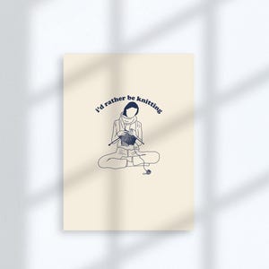 Pu&ograve; includere: Un biglietto beige con un disegno a linea blu di una persona seduta a gambe incrociate e che fa la maglia. Il testo "I'd rather be knitting" &egrave; scritto in blu, con un carattere curvo.