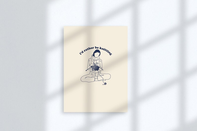 Pu&ograve; includere: Un biglietto beige con un disegno a linea blu di una persona seduta a gambe incrociate e che fa la maglia. Il testo "I'd rather be knitting" &egrave; scritto in blu, con un carattere curvo.