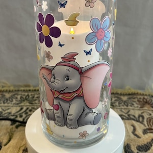 May include: A cylindrical glass candle holder featuring a graphic of a grey elephant with large pink ears, surrounded by colorful flowers, butterflies, and hearts. Inside the glass sits a white pillar candle with a glowing flame on a white base.