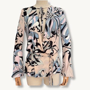 May include: A long-sleeved wrap blouse featuring a pale pink base with a bold, abstract floral pattern in black, light blue, and white. The garment includes a deep V-neckline with a tie closure at the neck and a wrap-around waist tie detail.