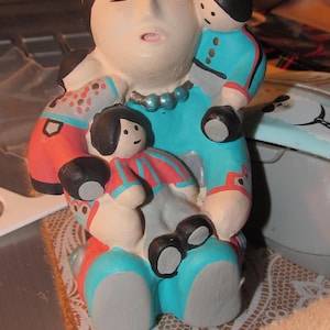 May include: A ceramic figurine of a woman with a red headband, holding two smaller figurines. The woman is wearing a turquoise and white dress with a beaded necklace. The smaller figurines are wearing blue and red clothing.