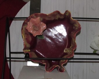 May include: A small, heart-shaped ceramic dish with a ruffled edge. The dish is a deep burgundy color with yellow accents and a pink rose decoration.