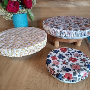 Puede incluir: Tres cubiertas de cuencos con estampados florales. Una es amarilla con un patr&oacute;n repetitivo, otra tiene un fondo blanco con flores rosas, naranjas y azules, y la tercera tiene un fondo blanco con flores rojas y azules. Las cubiertas est&aacute;n sobre cuencos y un peque&ntilde;o soporte de madera.