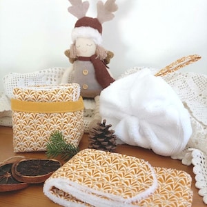 Puede incluir: Una cesta de tela y un paño plegado con un patrón de abanicos dorados y blancos, una esponja de baño de felpa blanca con un lazo estampado, una muñeca con astas de reno, una piña y dos rodajas de naranja seca sobre una superficie de madera.