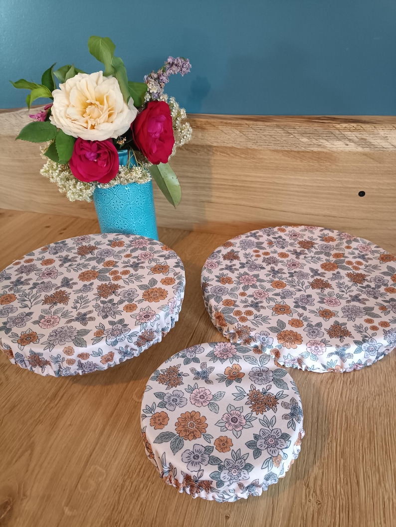 Puede incluir: Tres cubiertas reutilizables para alimentos con un estampado floral en tonos rosa, naranja y azul sobre un fondo blanco. Un peque&ntilde;o jarr&oacute;n turquesa contiene un ramo de flores con rosas blancas y rojas. Las cubiertas son de diferentes tama&ntilde;os.