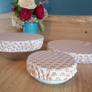 Puede incluir: Tres cuencos con cubiertas de tela. Las cubiertas tienen un fondo blanco con un patr&oacute;n repetido de dise&ntilde;os florales dorados y rojos. Los cuencos est&aacute;n sobre una superficie de madera, con un jarr&oacute;n de flores en el fondo.