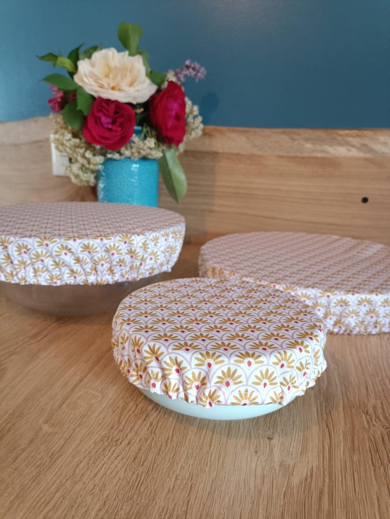 Puede incluir: Tres cuencos con cubiertas de tela. Las cubiertas tienen un fondo blanco con un patr&oacute;n repetido de dise&ntilde;os florales dorados y rojos. Los cuencos est&aacute;n sobre una superficie de madera, con un jarr&oacute;n de flores en el fondo.