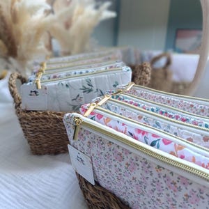 Peut inclure: Plusieurs trousses à crayons à motifs floraux avec des fermetures éclair dorées sont présentées dans un panier en osier. Les trousses présentent divers motifs floraux pastel sur fond blanc. Le panier est posé sur une surface blanche, avec un miroir et des fleurs séchées en arrière-plan.
