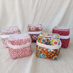 Puede incluir: Cinco cestas de tela pequeñas, cada una con una pila de almohadillas de tela cuadradas a juego. Las cestas presentan varios patrones, incluyendo estampado de leopardo rosa, diseños florales y un estampado de flores naranja y azul con borde blanco.