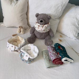 Puede incluir: Un oso de peluche gris con un calentador de cuello blanco con estampado floral, acompañado de dos calentadores de cuello adicionales, tres telas rectangulares plegadas con varios patrones y un par de zapatos de bebé color crema.