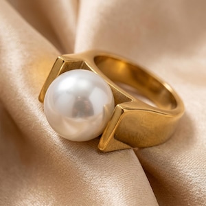 May include: A gold-colored ring with a large, round, white pearl. The ring has a modern design with geometric shapes. The pearl is centered and appears to be the focal point of the ring. The ring is set against a gold-colored fabric.