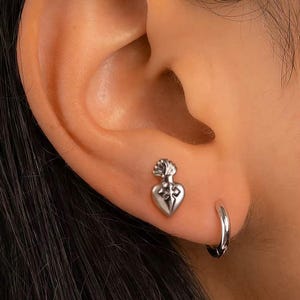May include: A silver-colored stud earring in the shape of a heart with a cross design. The earring is worn in the ear, next to a small silver hoop earring. The image is a close-up of the ear.