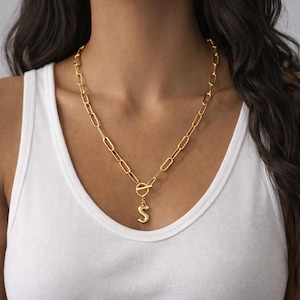 Op de afbeelding: Goudkleurige ketting met een ovaal schakelontwerp. De ketting heeft een ronde sluiting en een hanger in de vorm van de letter "S". De ketting wordt gedragen over een witte tanktop.