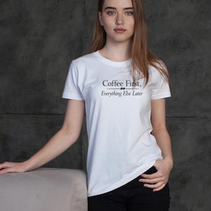 May include: White t-shirt with the text "Coffee First, Everything Else Later" in black. The shirt has short sleeves and a crew neck. The model is wearing black pants.