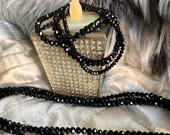 May include: A black beaded necklace draped over a silver, crystal-studded candle holder with a faux candle. The necklace is composed of multiple strands of small, faceted beads. The background features a soft, gray faux fur.