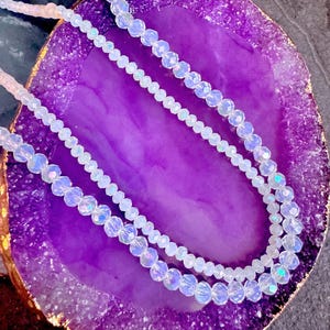 May include: Two delicate necklaces with iridescent, faceted beads. The necklaces are displayed on a vibrant purple geode slice with a gold-colored edge. The beads reflect light, creating a shimmering effect. The necklaces are likely intended for fashion or jewelry.