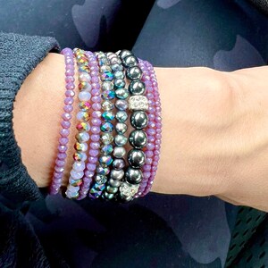 May include: A collection of beaded bracelets in various styles and colors. The bracelets include shades of purple, iridescent, and dark gray beads. Some bracelets feature crystal accents, creating a layered look.