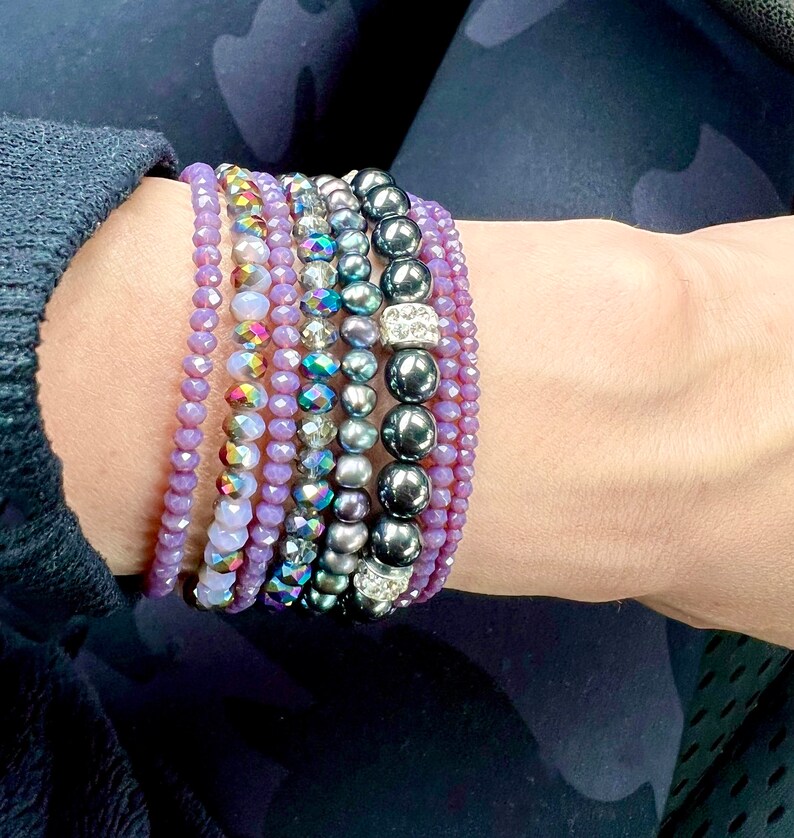 May include: A collection of beaded bracelets in various styles and colors. The bracelets include shades of purple, iridescent, and dark gray beads. Some bracelets feature crystal accents, creating a layered look.