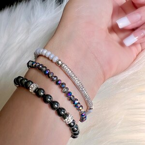 May include: Three beaded bracelets in varying styles. The top bracelet features light gray beads. The middle bracelet has iridescent, dark gray beads and a silver bar with clear crystals. The bottom bracelet has black beads with silver and crystal accents.