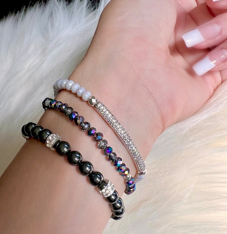 May include: Three beaded bracelets in varying styles. The top bracelet features light gray beads. The middle bracelet has iridescent, dark gray beads and a silver bar with clear crystals. The bottom bracelet has black beads with silver and crystal accents.