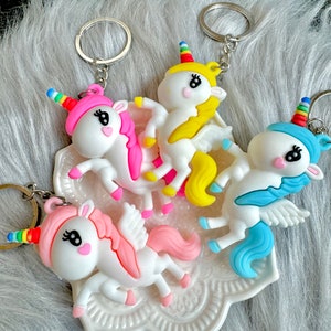May include: Four unicorn keychains with rainbow horns, pink, yellow, and blue manes and wings. The keychains are white with black eyes and are attached to silver keyrings. The unicorns are on a white, decorative, scalloped dish.