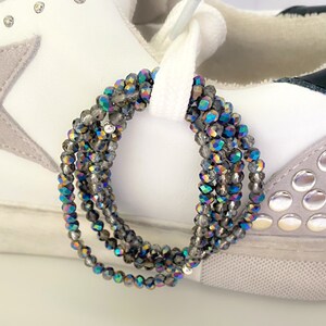 May include: Close-up of a white sneaker with gray suede accents and silver studs. Strands of iridescent gray beads are looped through the white shoelaces, adding a touch of sparkle and color to the shoe.