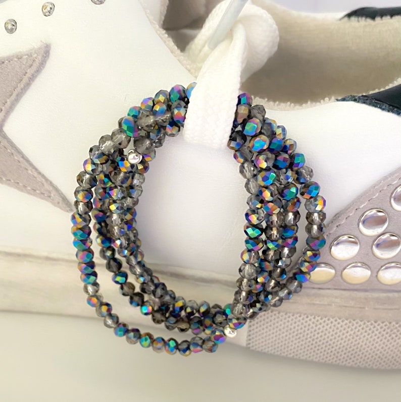 May include: Close-up of a white sneaker with gray suede accents and silver studs. Strands of iridescent gray beads are looped through the white shoelaces, adding a touch of sparkle and color to the shoe.