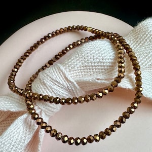 May include: Two strands of bronze-colored beaded jewelry. The beads are small, faceted, and strung together. The jewelry is resting on a white fabric and a pink surface. The beads have a metallic sheen.