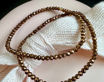 May include: Two strands of bronze-colored beaded jewelry. The beads are small, faceted, and strung together. The jewelry is resting on a white fabric and a pink surface. The beads have a metallic sheen.